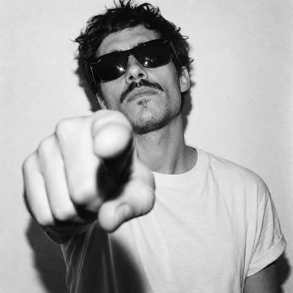 Retrato de busto de un serigrafista joven con gafas de sol negras y camiseta blanca, señalando directamente a la cámara con el dedo índice en primer plano, luz dura de flash frontal con sombras marcadas, fondo blanco liso, fotografía en blanco y negro de alto contraste